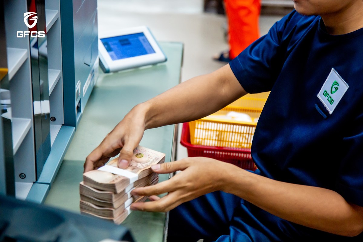 A GFCS cash-processing employee squares a strapped bundle of Thai banknotes at a counting station.
